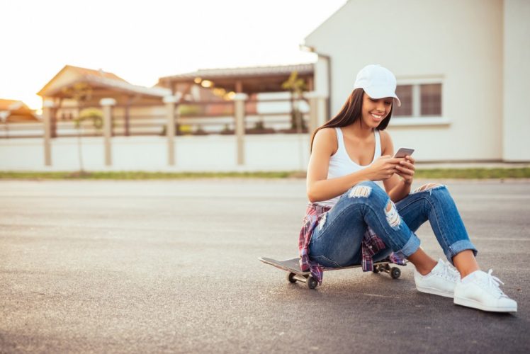 Woman sitting on a skateboard and texting on a mobile phone.