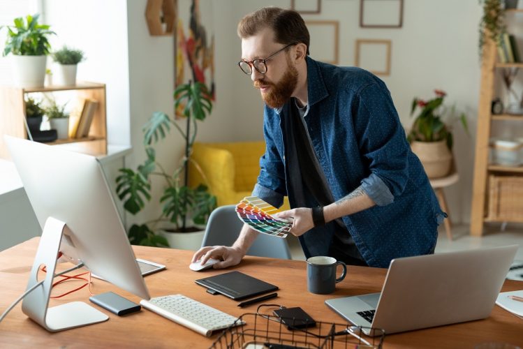 Young creative software developer or web designer bending over wooden table in front of computer monitor while choosing color from palette